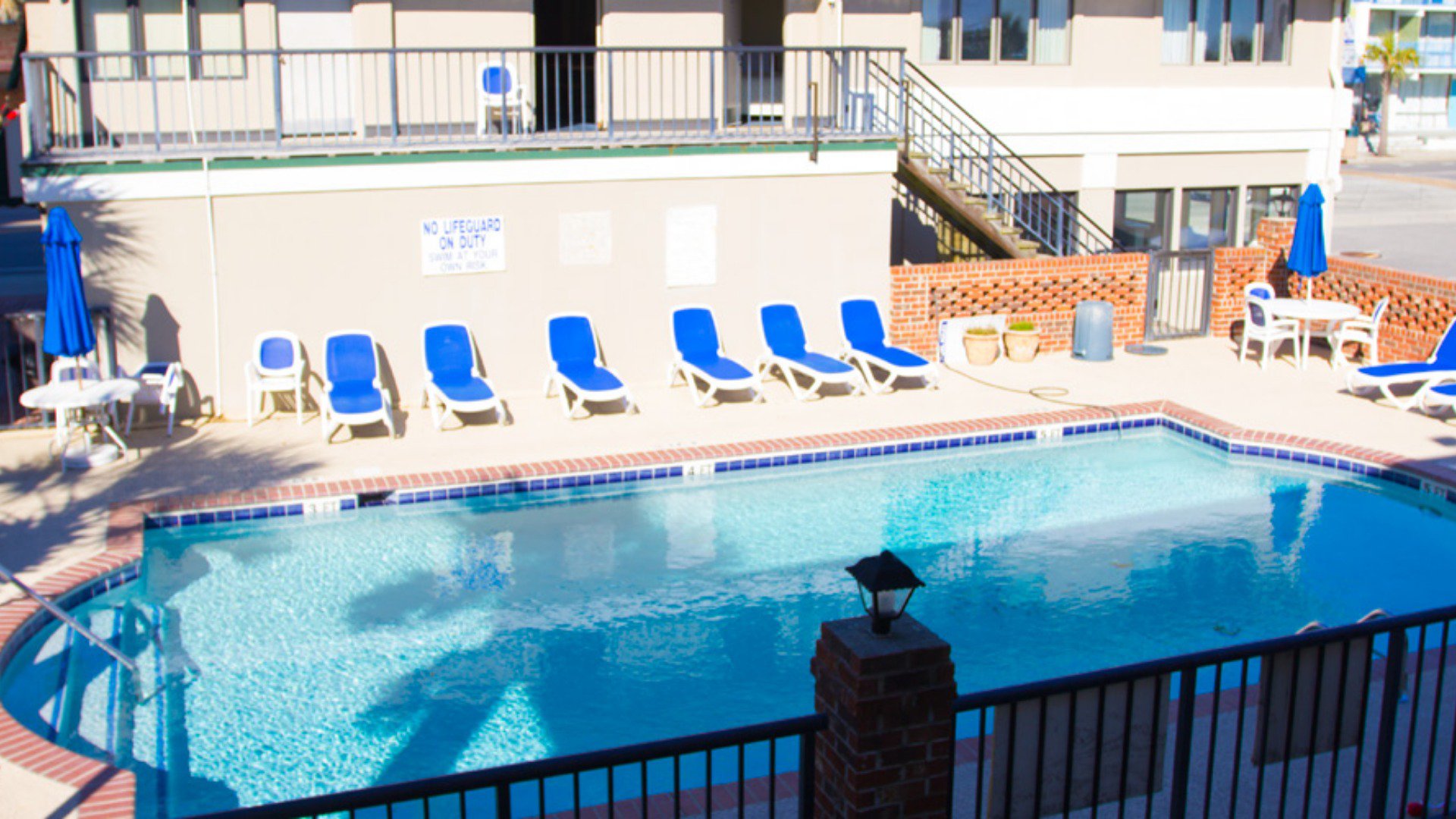 A swimming pool with blue water and red brick surroundings, surrounded by blue lounge chairs, white tables, and blue umbrellas.