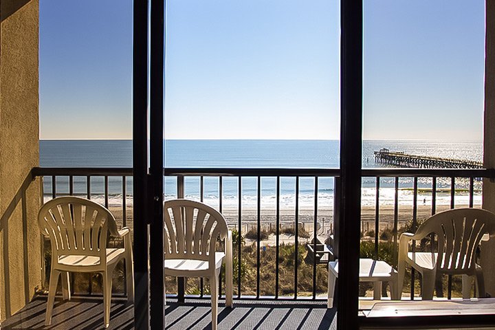A balcony with white plastic chairs and a table overlooking the ocean and a pier.